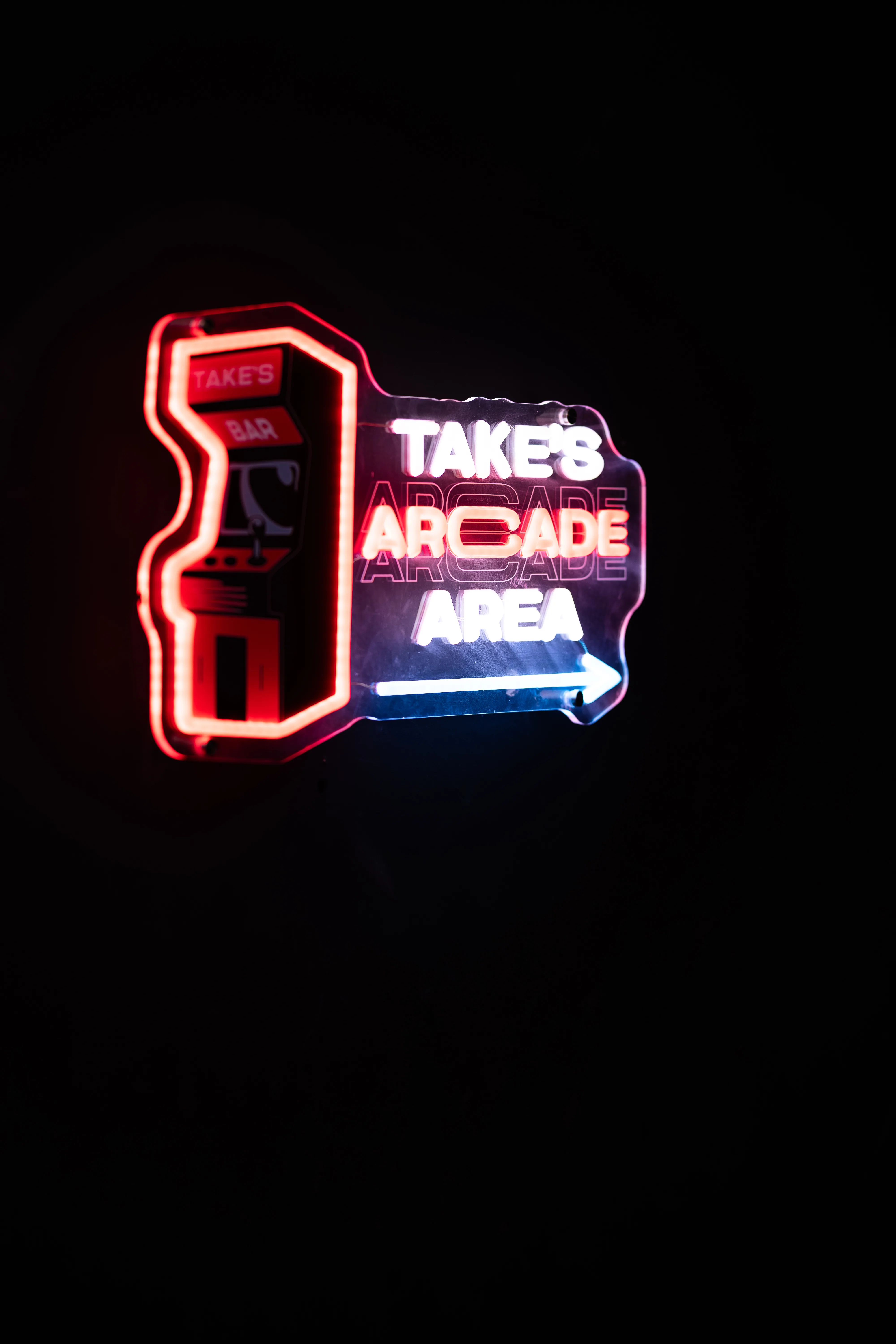 Ein leuchtendes Neon-Schild vor schwarzem Hintergrund in der Silhouette eines Arcade-Spielautomaten. In rot-weißer Leuchtschrift steht dort TAKE'S ARCADE AREA mit einem Pfeil, der nach rechts weist.