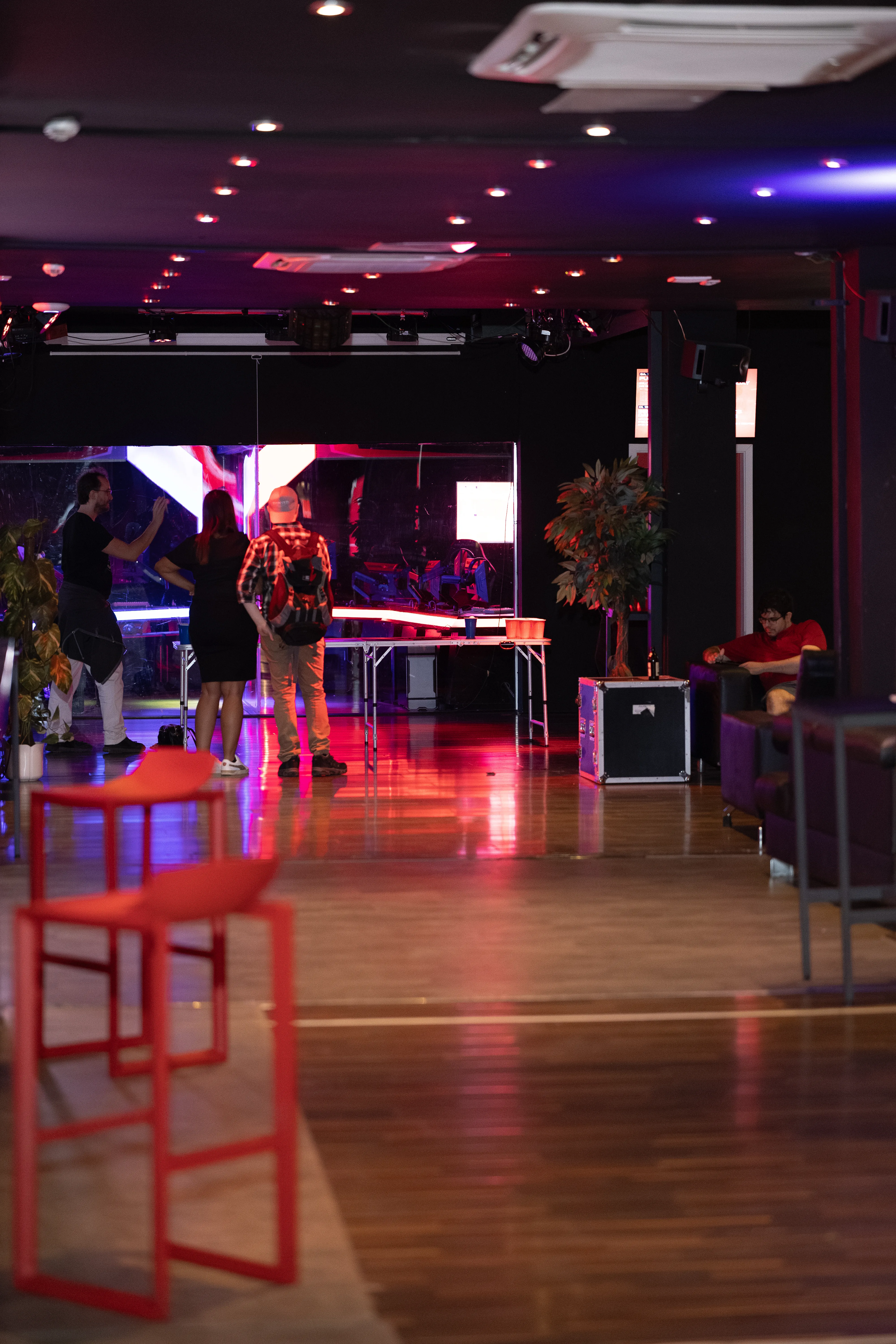 Ein schummrig rot beleuchteter Raum einer Event-Location. Im Vordergrund stehen rote Barhocker, während im Hintergrund Leute an einem Klapptisch entspannt eine Runde Beer Pong spielen.