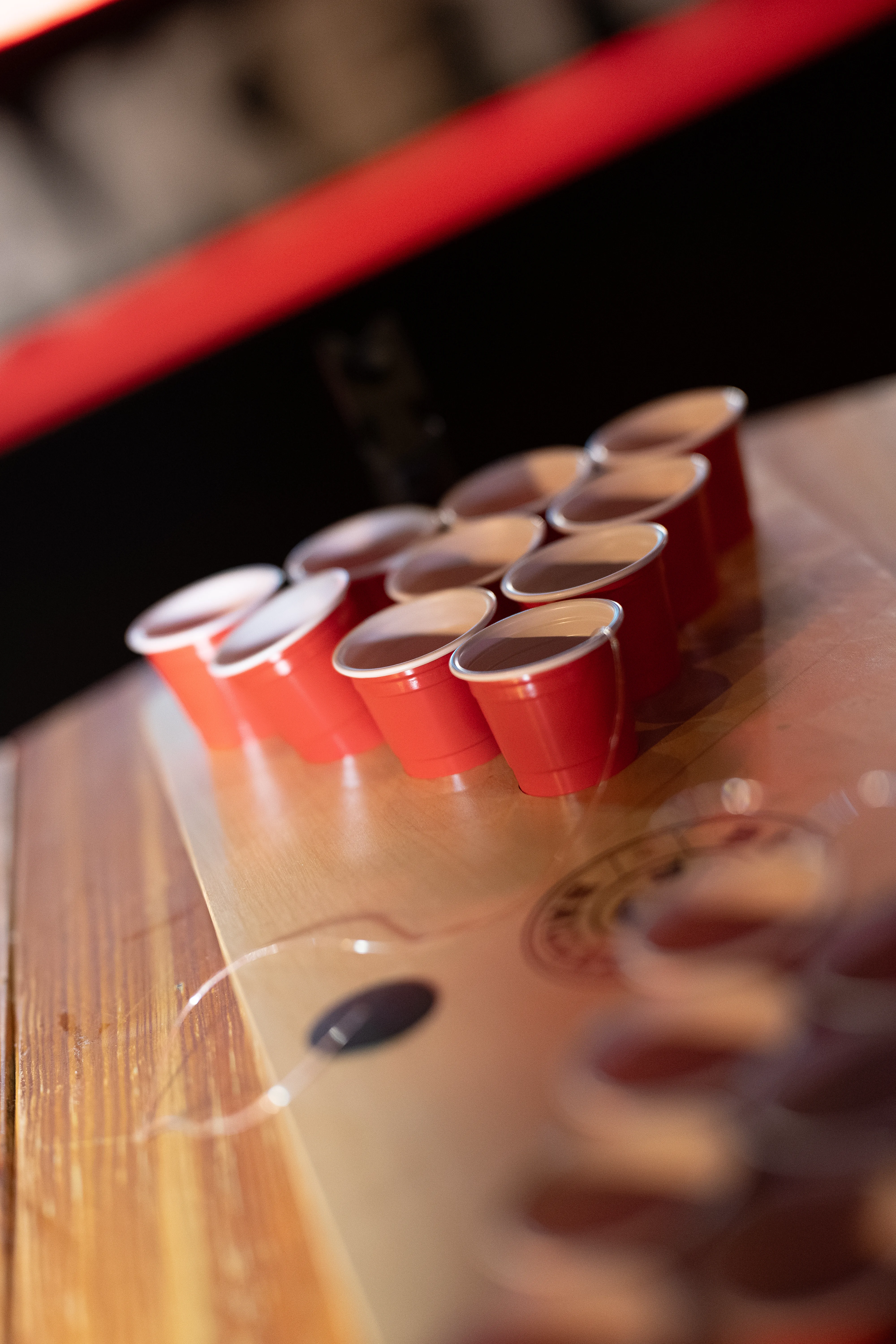 Nahaufnahme eines Beer-Pong-Spiels: Rote Plastikbecher stehen dicht gedrängt in einer Dreiecksformation auf einem Holztisch. Der Hintergrund und der unmittelbare Vordergrund verschwinden in einer weichen Unschärfe.