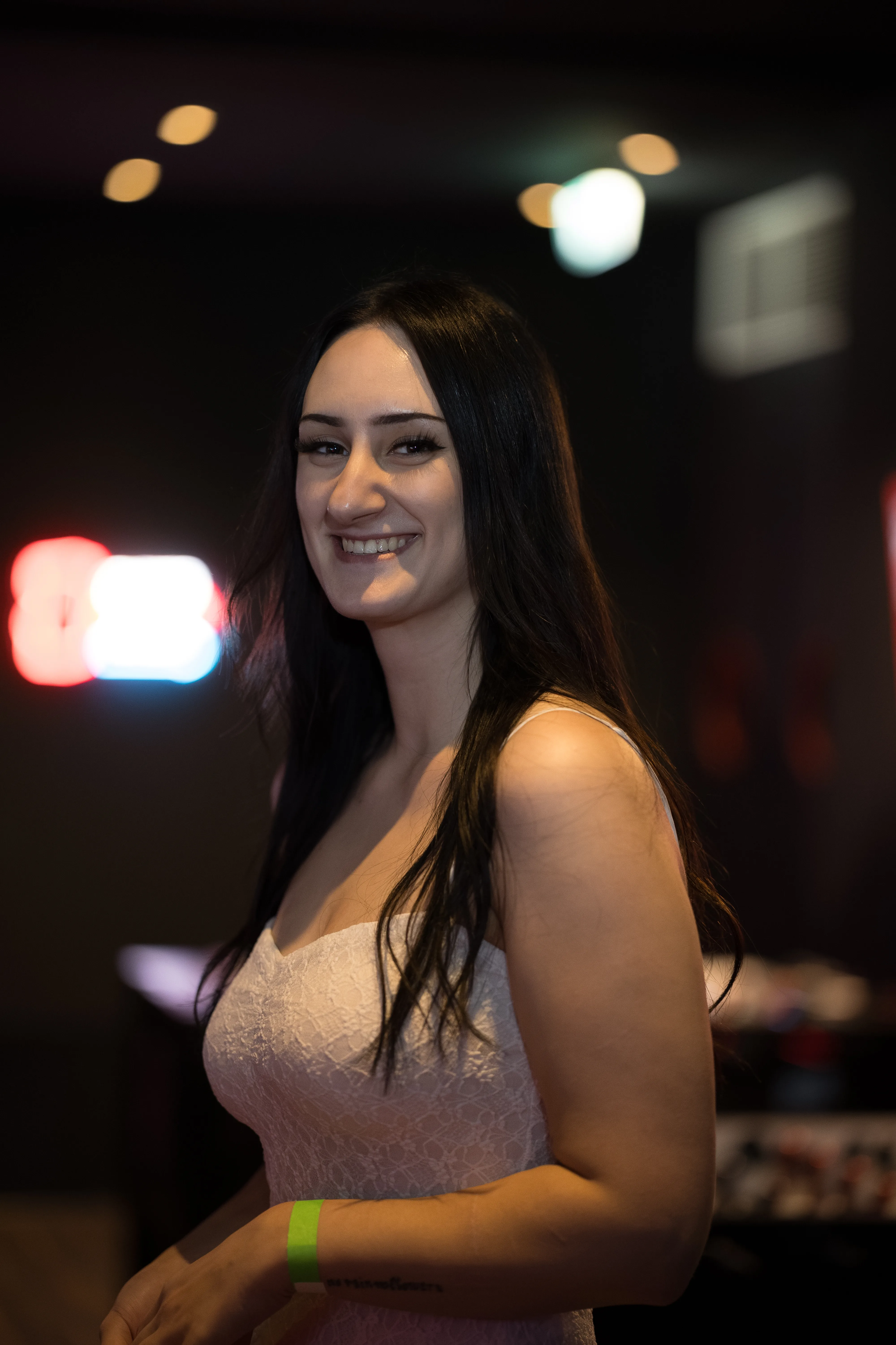 Eine lächelnde junge Frau mit langen dunklen Haaren trägt ein weißes Spitzenkleid und ein grünes Event-Bändchen am Handgelenk. Sie steht in einer schummrig beleuchteten Location mit unscharfen Neonlichtern im Hintergrund.