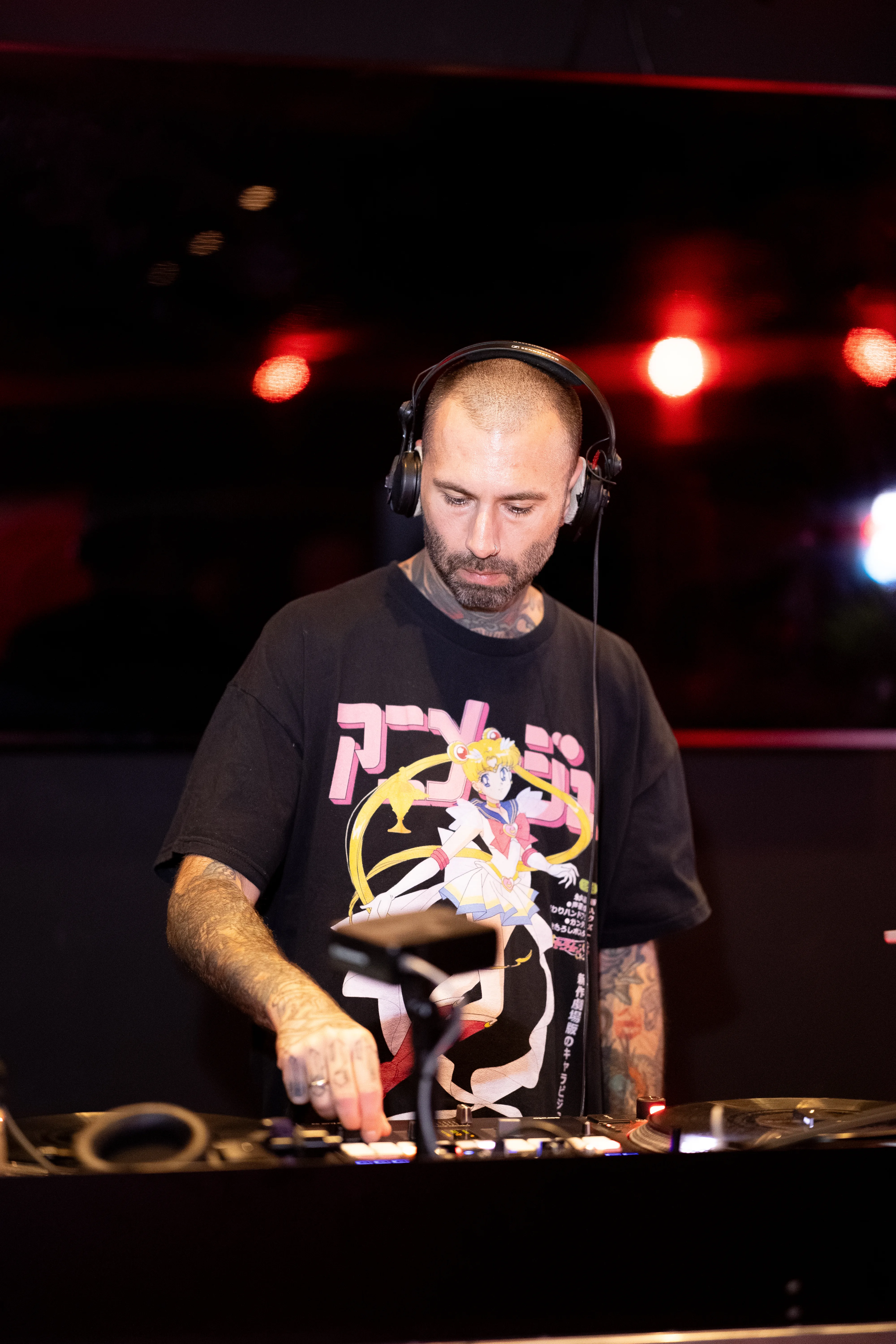 Ein tätowierter DJ mit kurzen Haaren und großen Kopfhörern steht konzentriert an seinem Mischpult. Er trägt ein schwarzes T-Shirt mit einem auffälligen Sailor-Moon-Aufdruck. Im Hintergrund leuchten rote Scheinwerfer.
