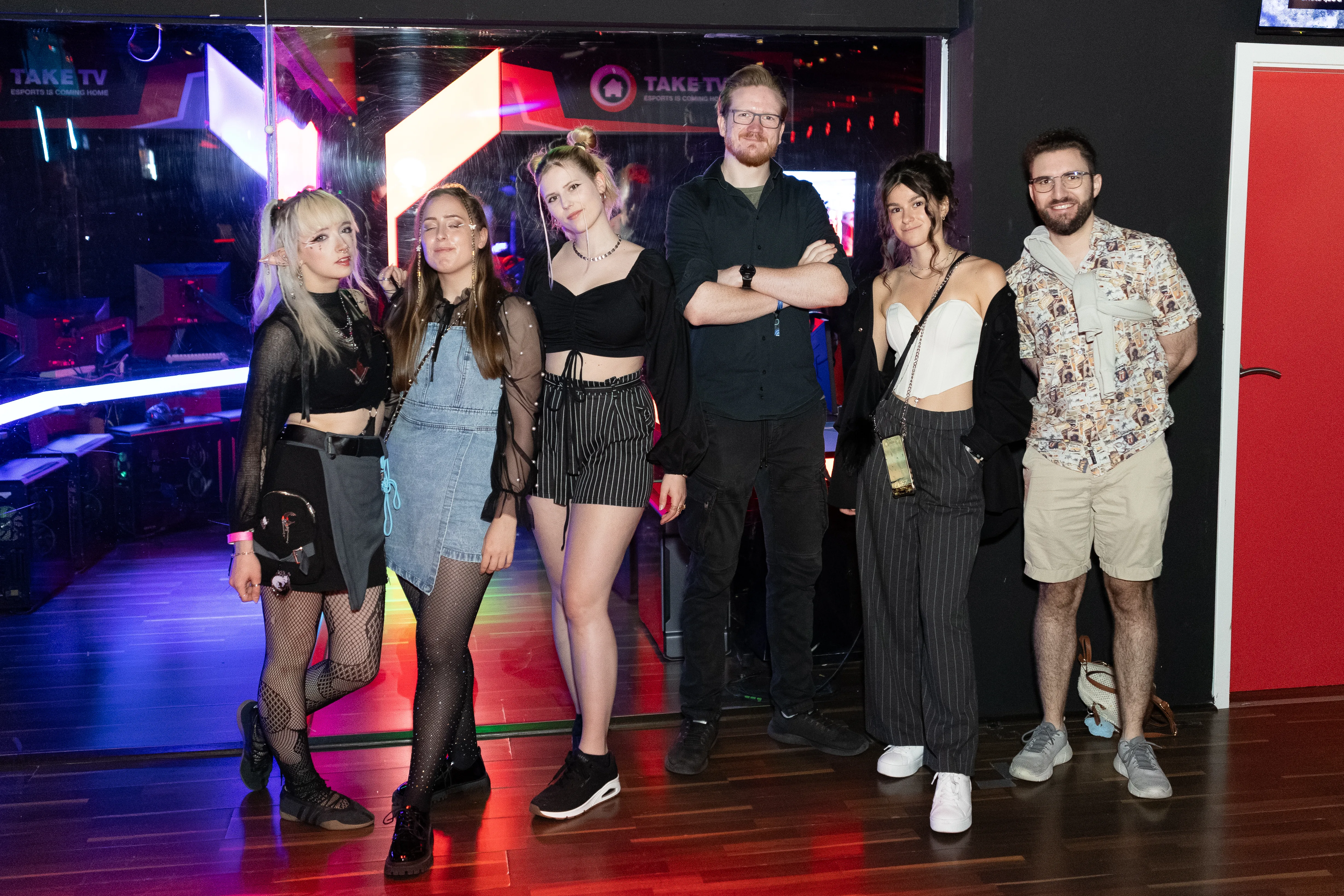 Eine Gruppe von sechs jungen Erwachsenen in individuellen Party-Outfits posiert fröhlich nebeneinander für ein Gruppenfoto in einer Event-Location. Hinter ihnen spiegelt sich buntes Licht in einer Glasscheibe mit einem TAKE TV-Logo.