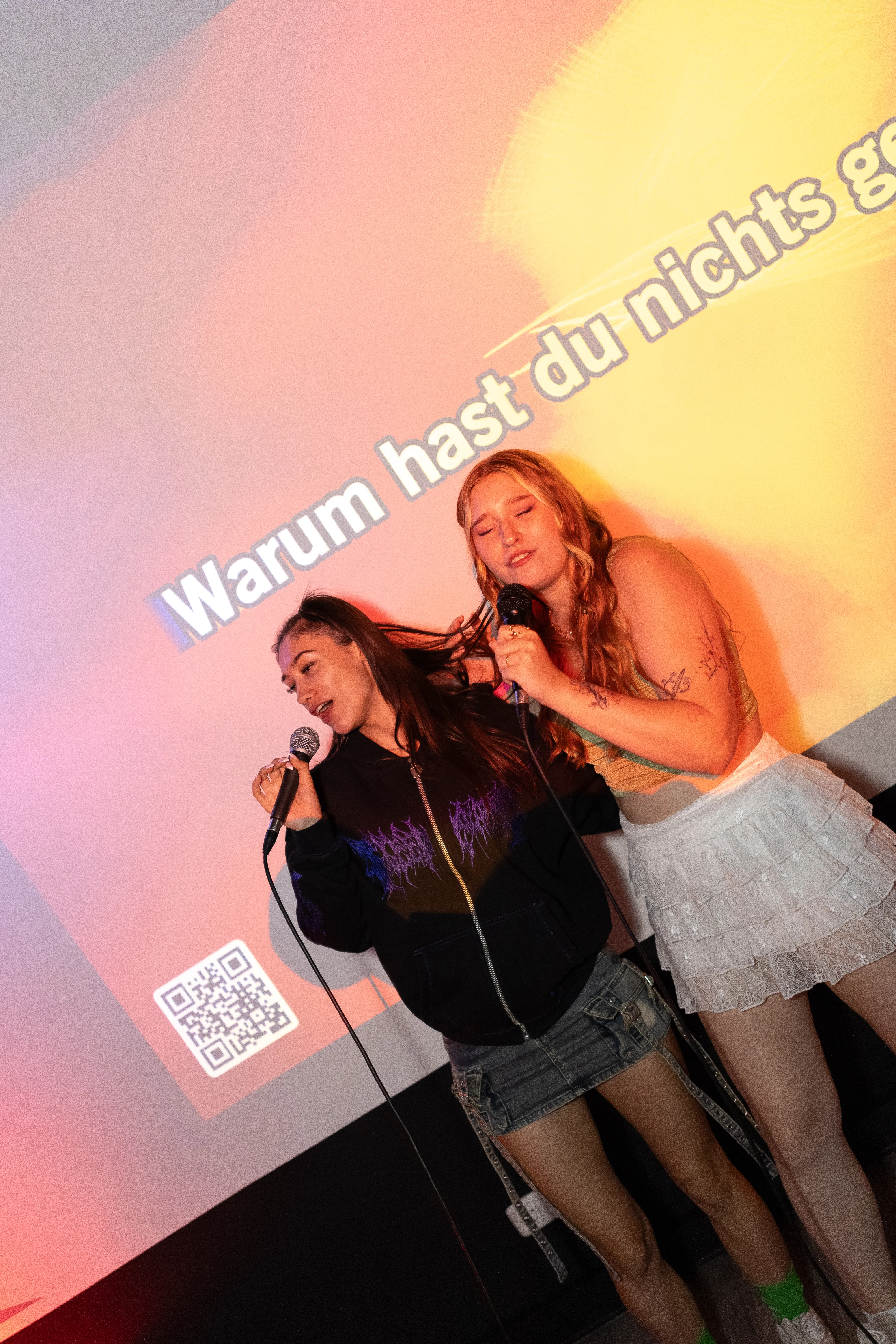 Zwei junge Frauen singen enthusiastisch in Mikrofone, während sie dicht nebeneinander stehen. Im Hintergrund leuchtet eine Projektion mit dem unvollständigen Text Warum hast du nichts g... und einem QR-Code.