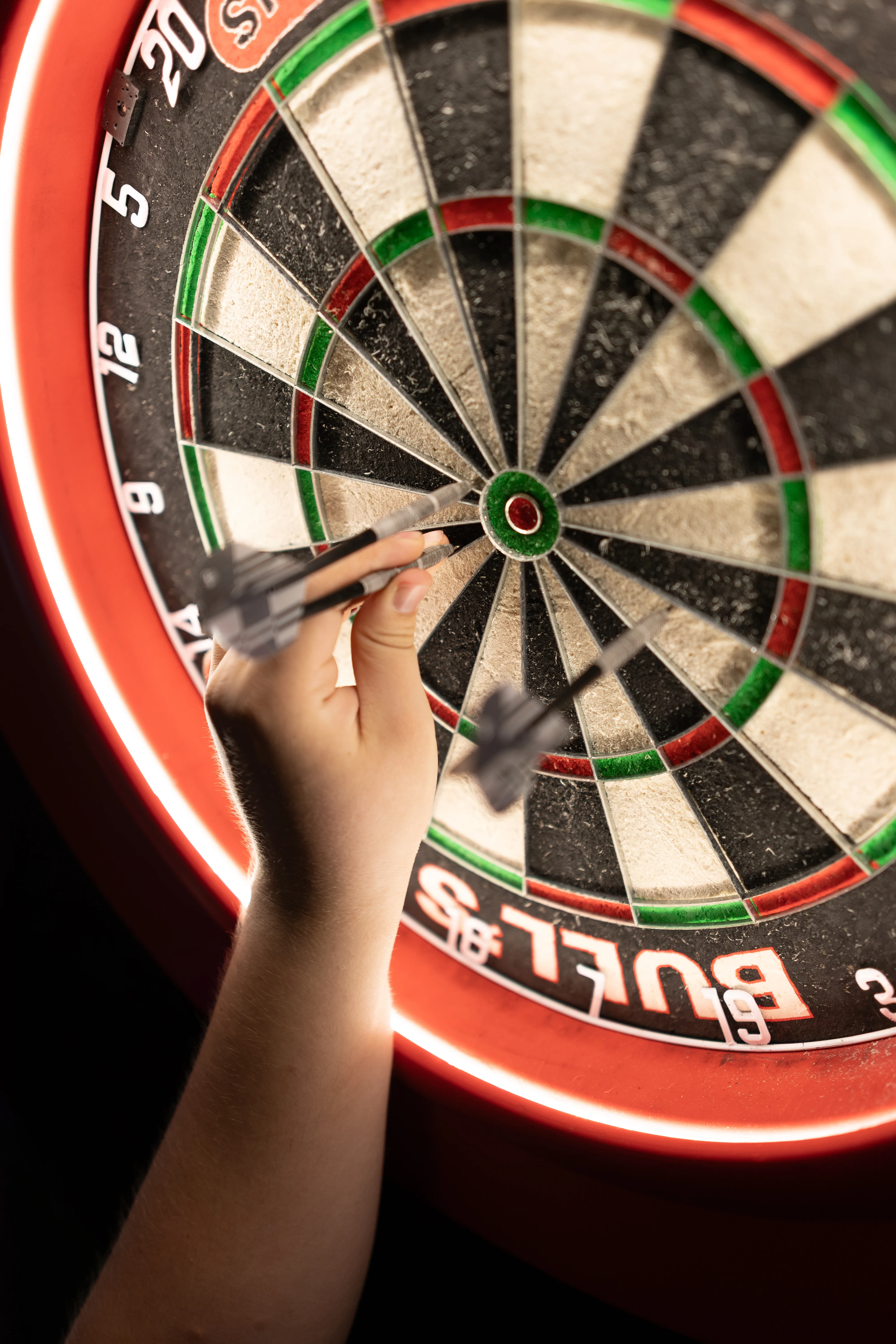 Nahaufnahme einer Hand, die zwei Dartpfeile hält, deren Spitzen direkt auf die Mitte (das Bullseye) einer Dartscheibe gerichtet sind. Die Dartscheibe wird von einem roten LED-Ring beleuchtet.