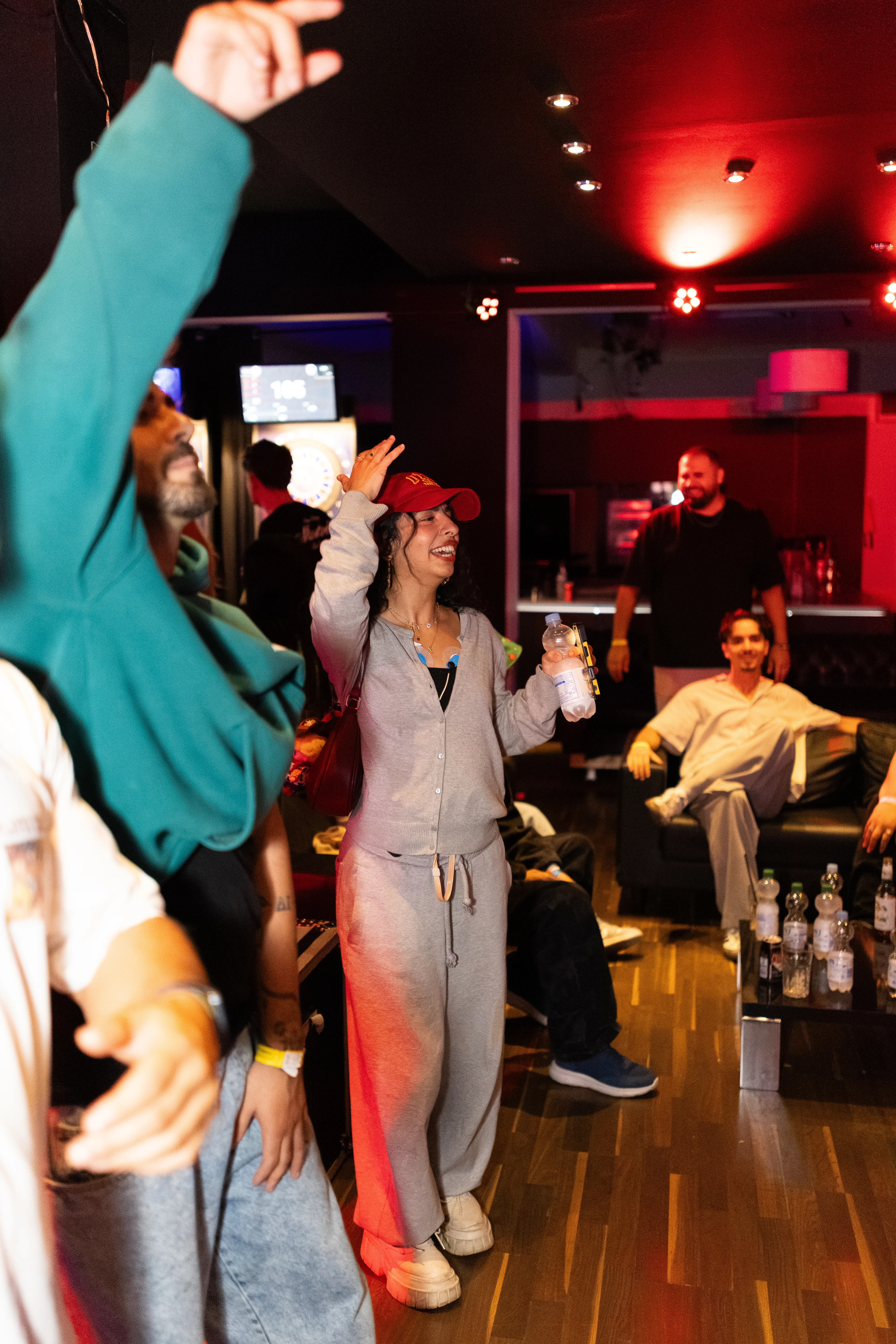 Eine fröhlich lachende junge Frau in einem grauen Zweiteiler und mit roter Kappe tanzt mit einer Wasserflasche in der Hand auf einer Party. Um sie herum sind weitere feiernde Personen in einer schummrig rot beleuchteten Location.