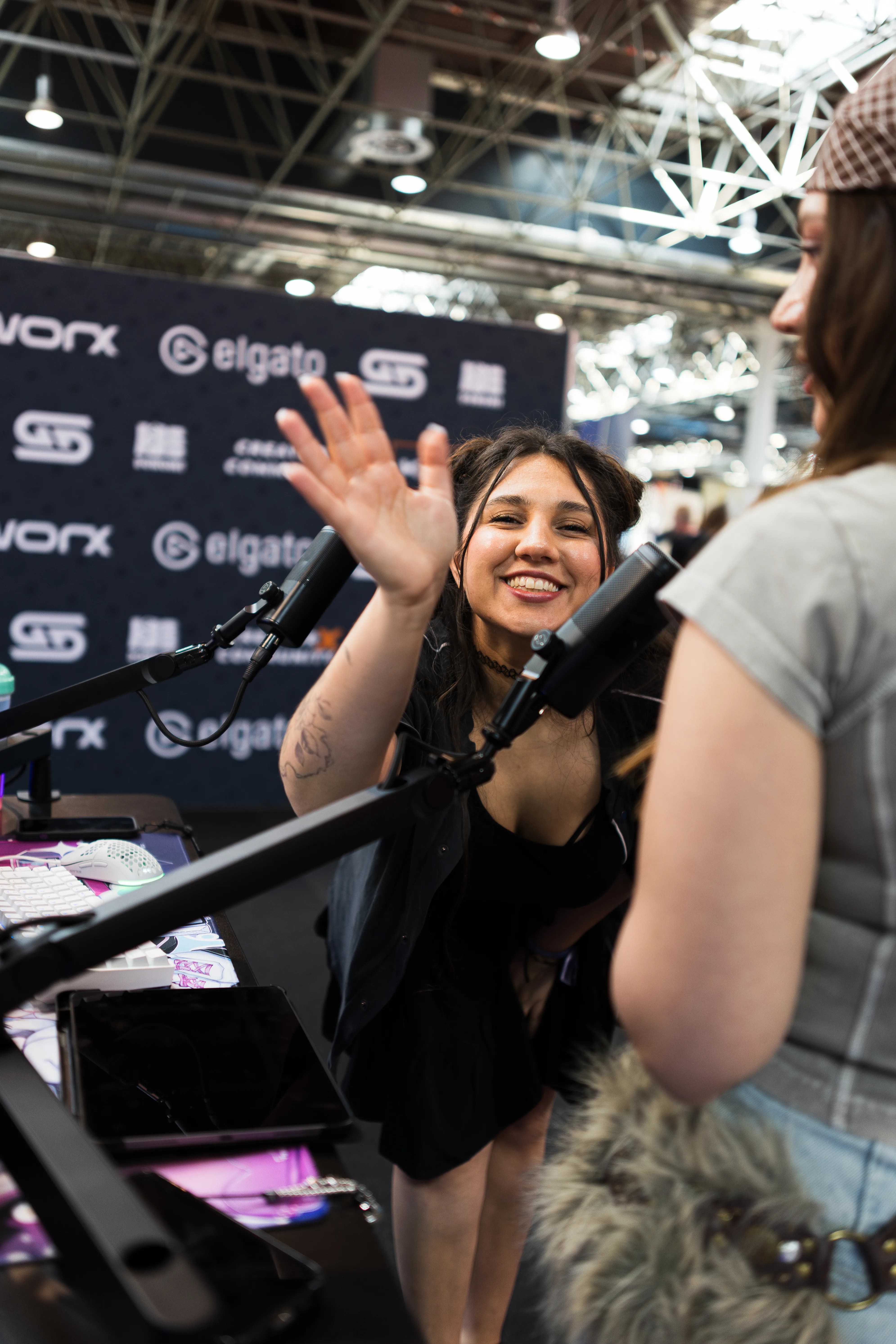 Eine freudig strahlende Frau mit dunklen Haaren beugt sich an einem Messestand zu einem Mikrofonarm nach vorne und winkt enthusiastisch mit der Hand. Eine weitere Person steht rechts mit dem Rücken zur Kamera im Bild.