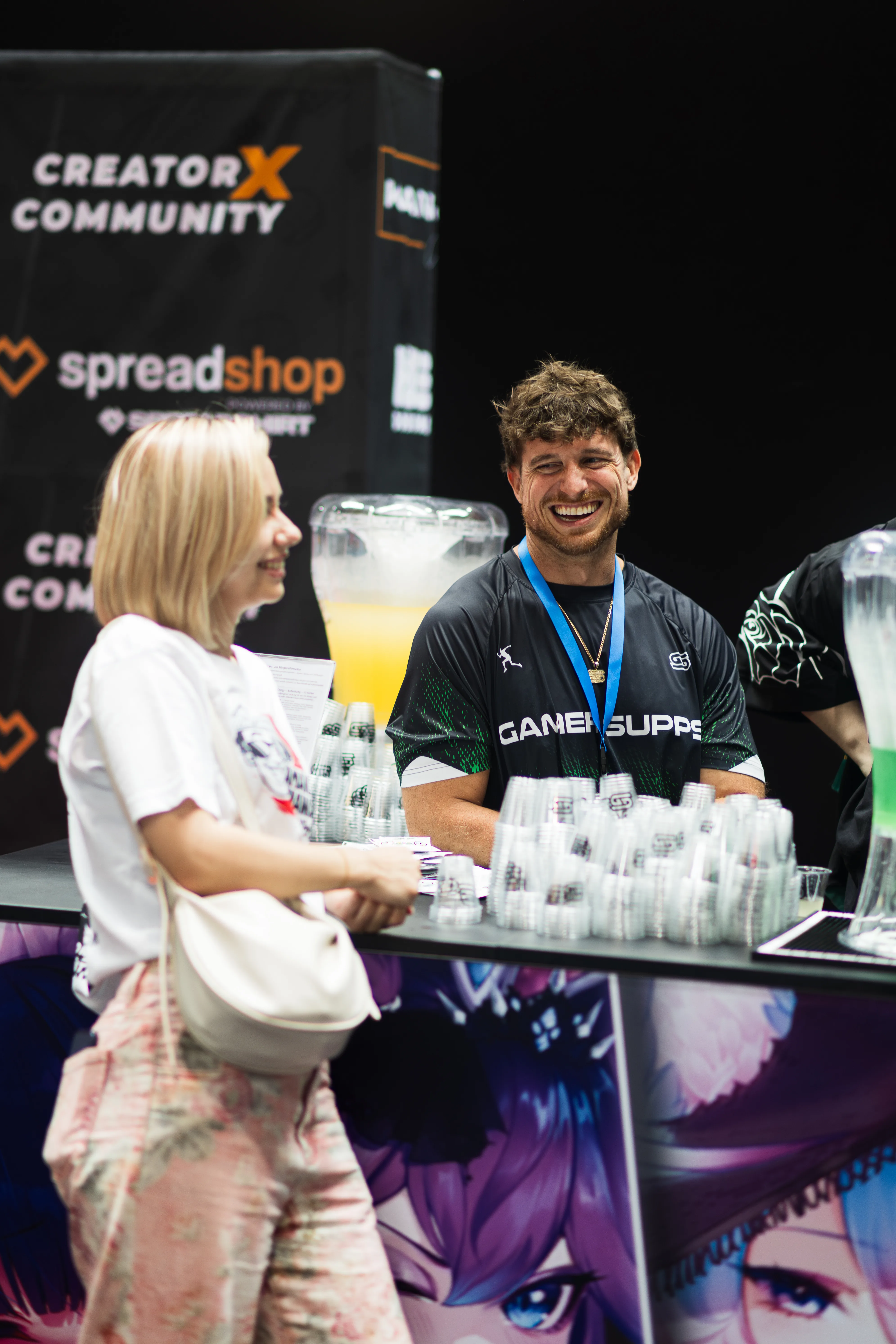 Ein fröhlich lächelnder Mann in einem schwarzen Gamersupps-T-Shirt steht an einem Messestand hinter einem Tresen mit Getränkespendern und vielen Bechern. Davor steht eine Frau mit blonden Haaren, die sich mit ihm unterhält. Im Hintergrund ist ein Banner mit Spreadshop-Logo zu sehen.