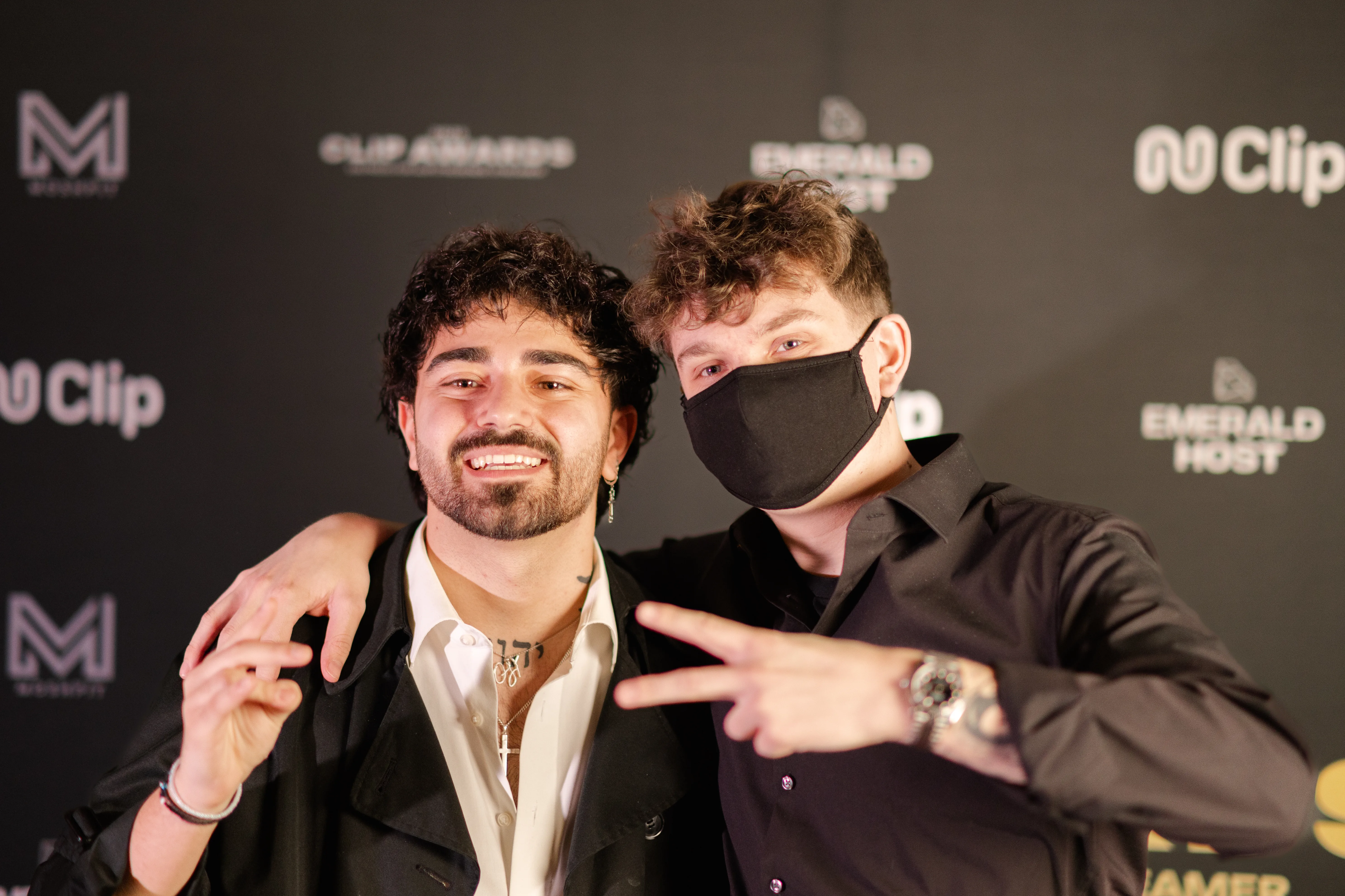 Zwei Männer posieren vor einer schwarzen Sponsorenwand: Der linke Mann mit dunklen Locken und Bart hat den Arm freundschaftlich um den rechten Mann mit schwarzem Hemd und schwarzer Gesichtsmaske gelegt, welcher ein Peace-Zeichen in die Kamera zeigt.