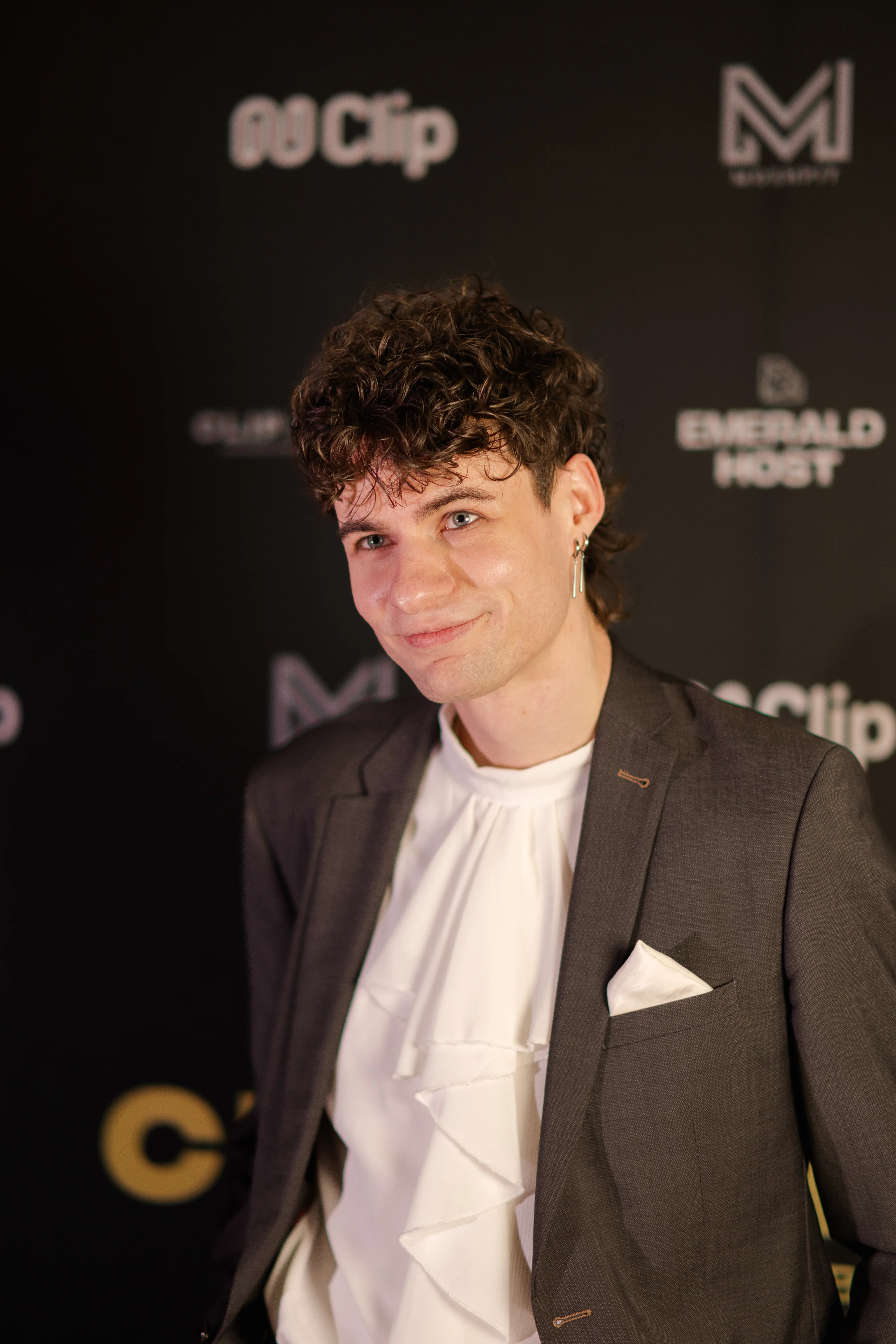Ein junger Mann mit lockigen braunen Haaren, der ein graues Sakko über einem weißen Hemd mit Rüschen trägt und leicht lächelnd vor einer schwarzen Fotowand der Clip Awards steht.