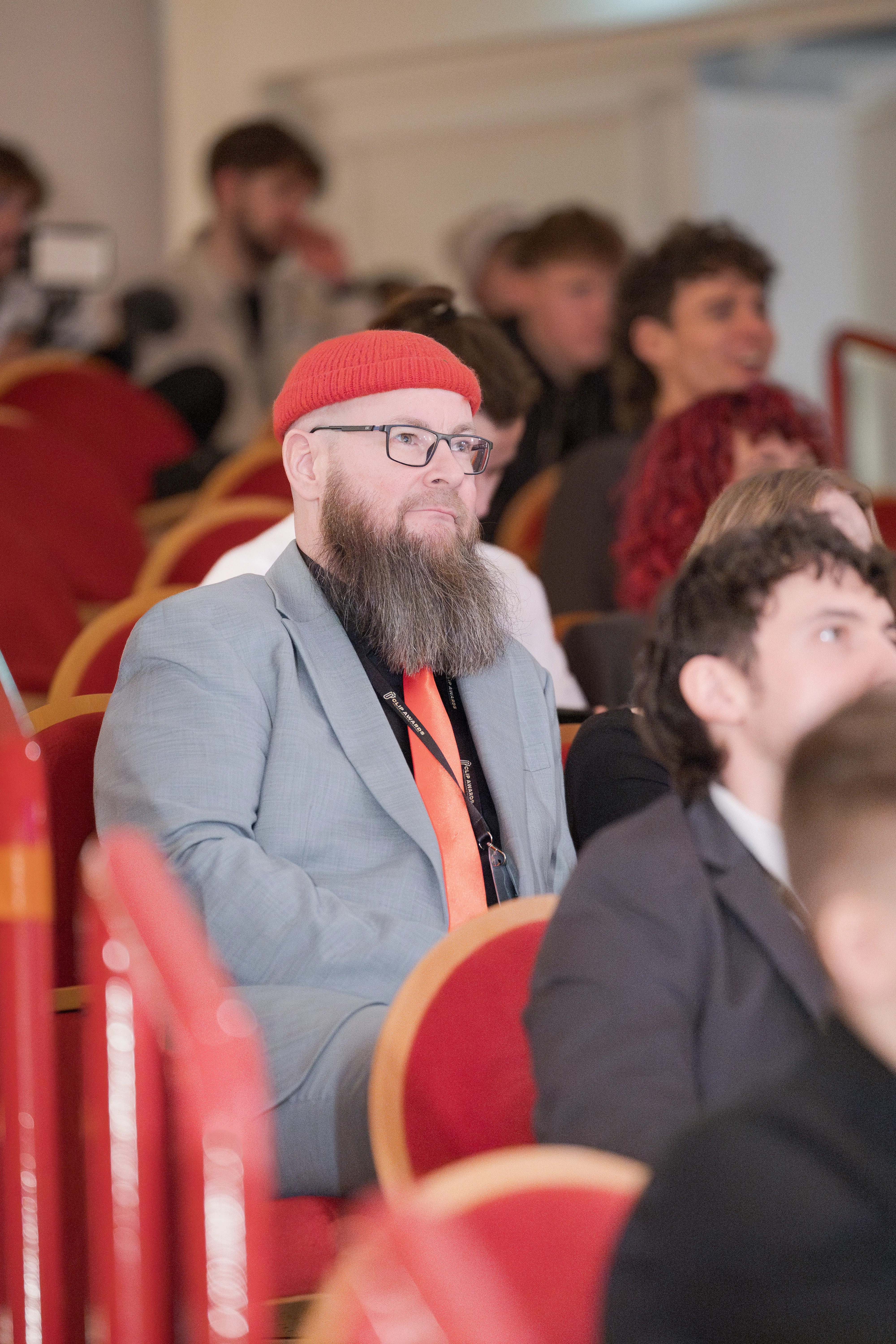 Ein Mann mit dichtem Vollbart, Brille, roter Mütze, grauem Anzug und leuchtend roter Krawatte sitzt aufmerksam auf einem roten Theatersitz im Publikum.