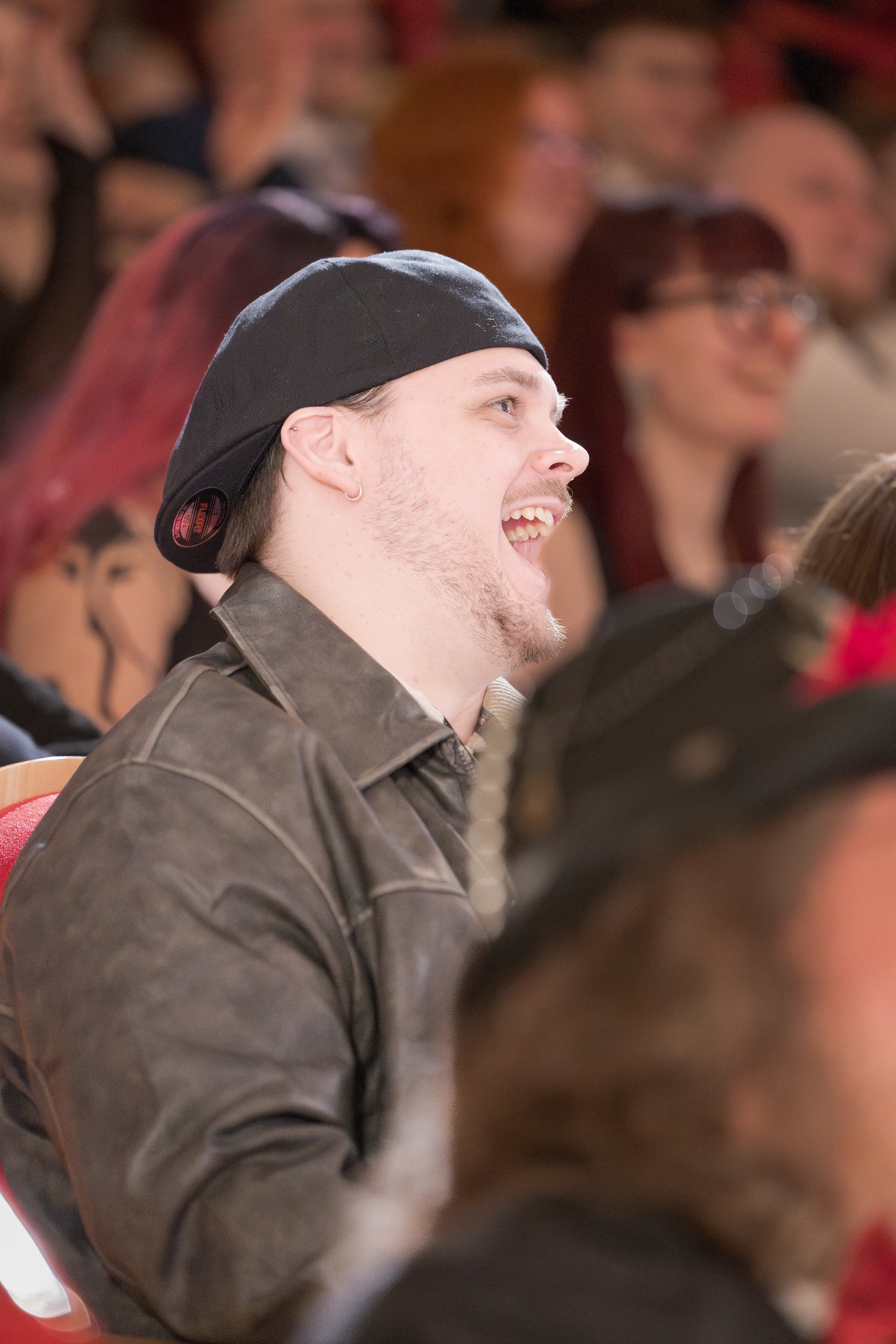 Ein fröhlich lachender Mann mit einem umgedrehten dunklen Cap und einer grauen Jacke in Lederoptik sitzt im Publikum.