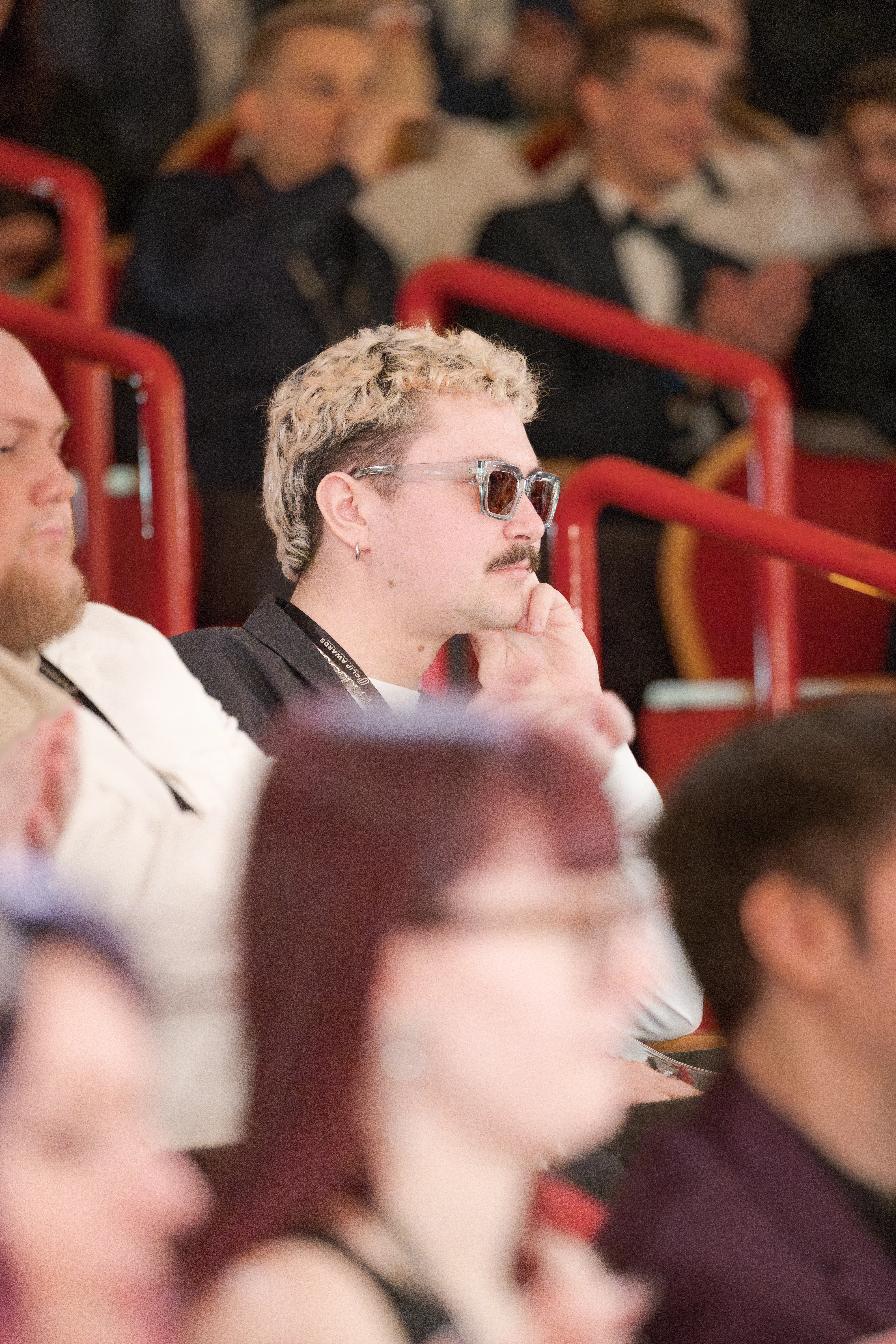 Eine Person mit hellblond gelocktem Vokuhila, Sonnenbrille und Schnurrbart sitzt nachdenklich mit aufgestütztem Kinn im Publikum.