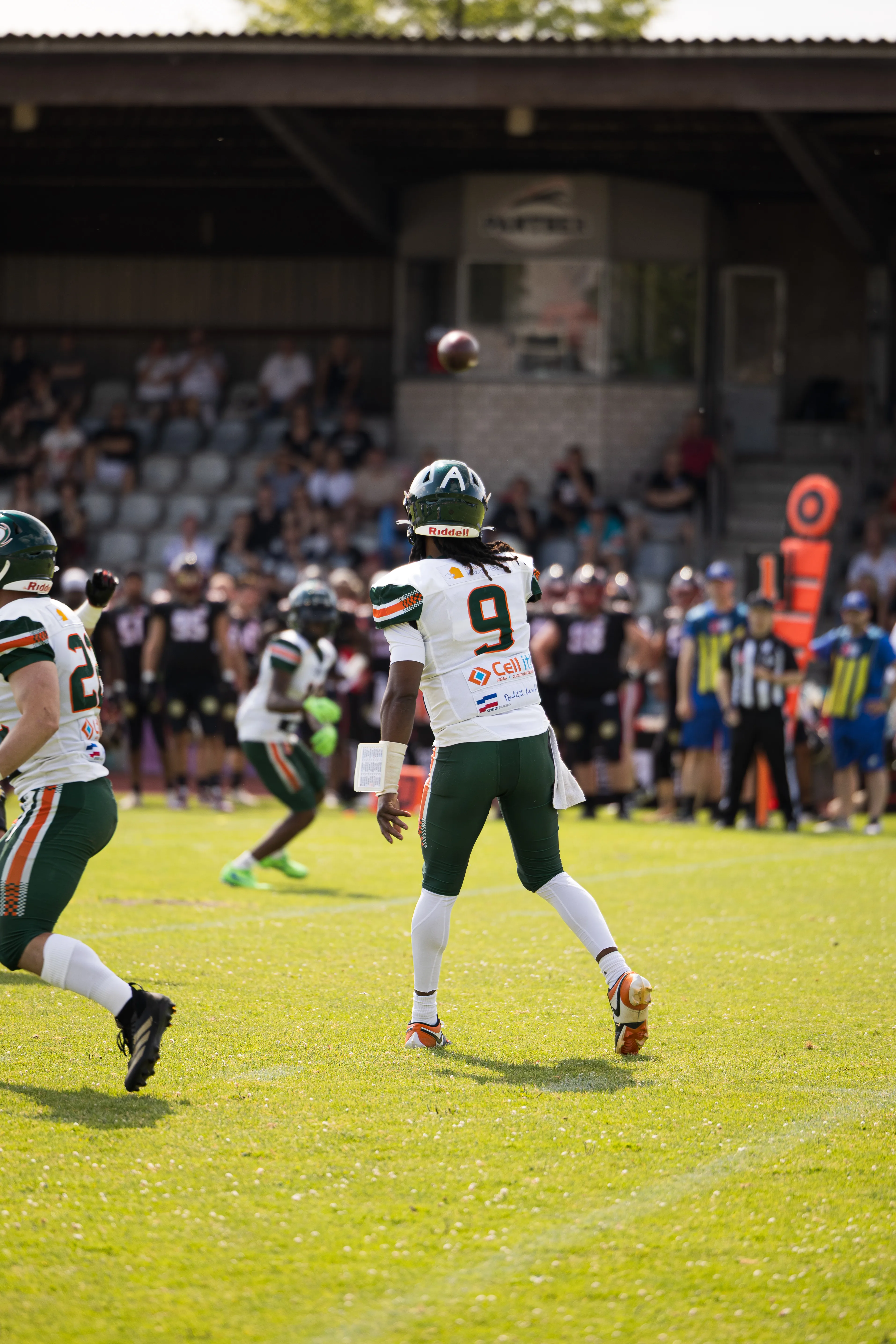 Ein American-Football-Spieler in einem weißen Trikot mit der Nummer 9 und grünem Helm ist in der Rückansicht zu sehen, wie er auf einem Rasenplatz einen Pass wirft. Der Football fliegt gerade durch die Luft vor einer unscharfen Tribüne mit Zuschauern.