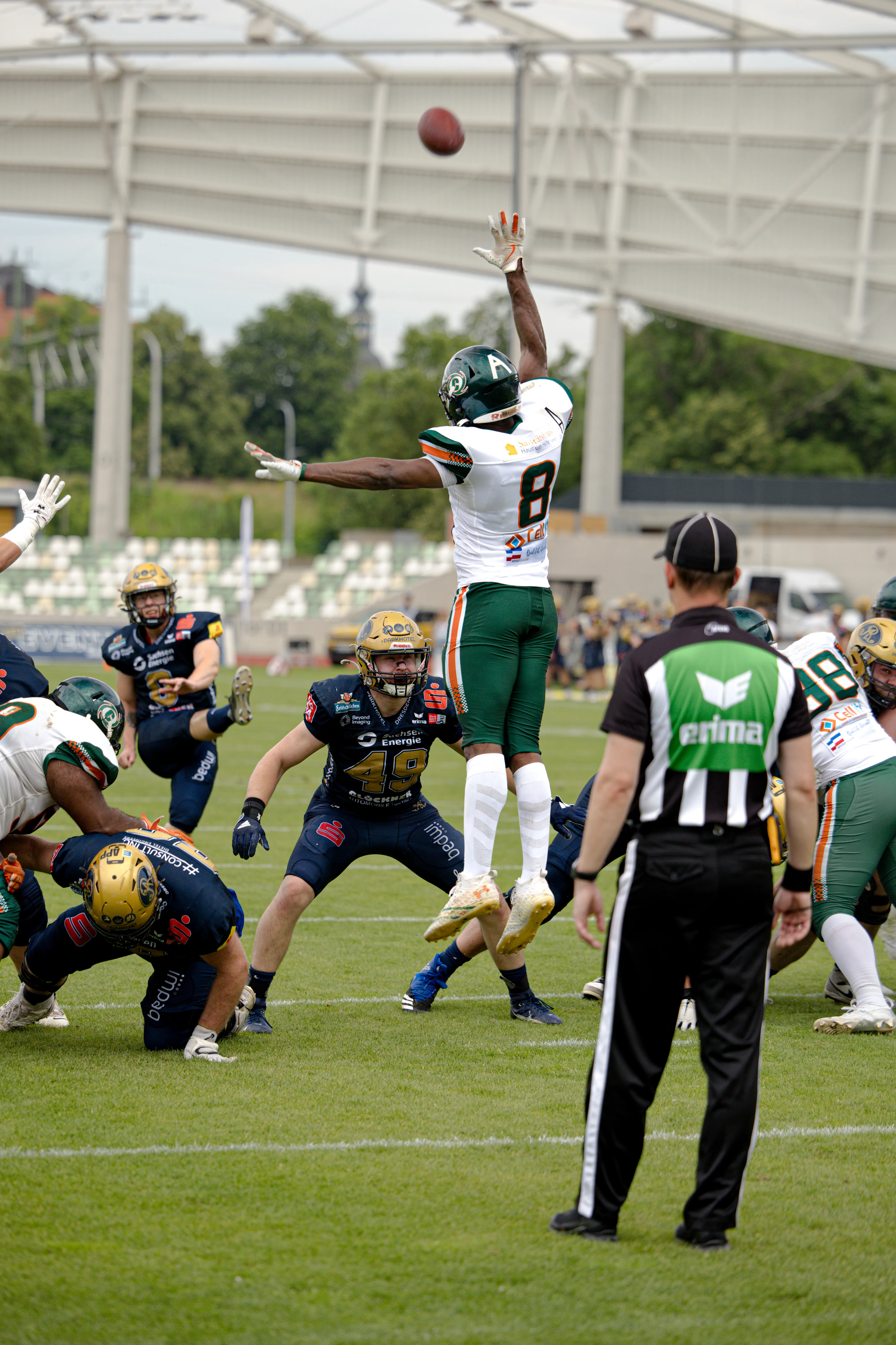 Ein Football-Spieler im weißen Trikot mit der Nummer 8 springt auf dem Spielfeld hoch in die Luft und streckt einen Arm aus, um einen herannahenden Football zu fangen. Um ihn herum stehen andere Spieler und ein Schiedsrichter.