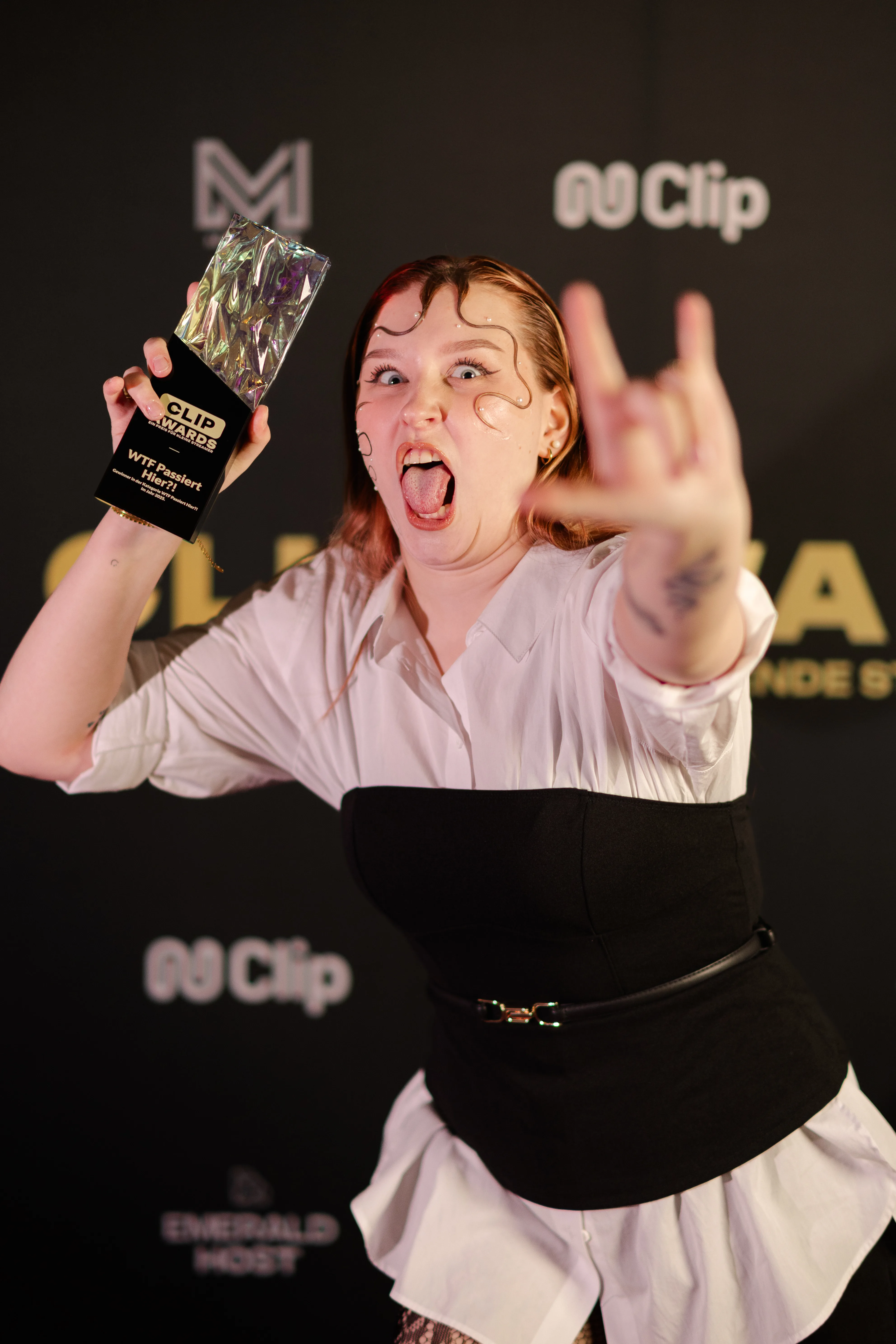 Eine junge Frau mit auffällig gestylten nassen Pony-Strähnen hält einen Clip Awards-Pokal hoch, streckt die Zunge heraus und zeigt mit der anderen Hand das Rock-on-Zeichen vor einer schwarzen Sponsorenwand.