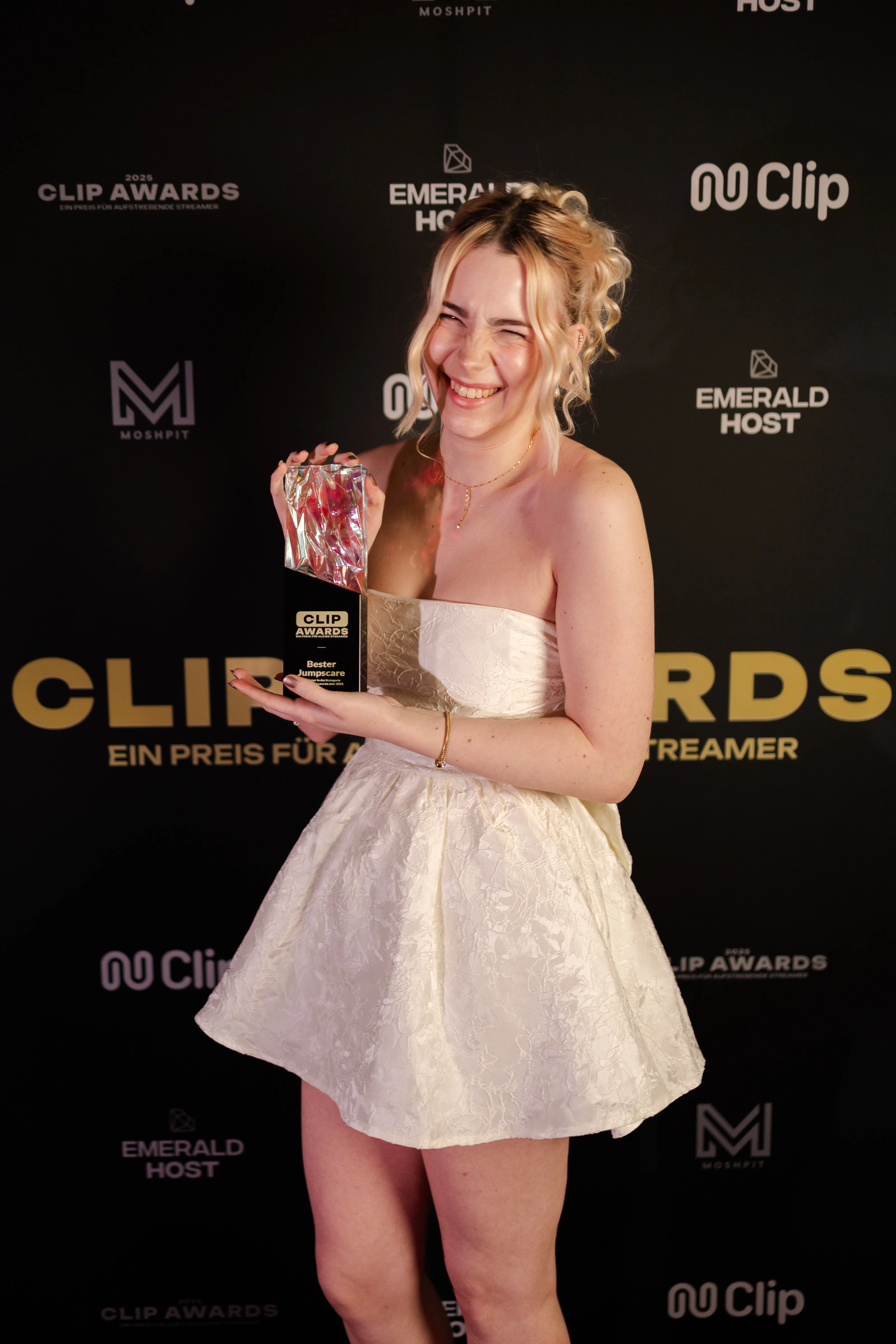Eine strahlende junge Frau mit blonden Haaren in einem kurzen, trägerlosen weißen Kleid hält freudig einen Clip Awards-Pokal vor einer schwarzen Sponsorenwand.