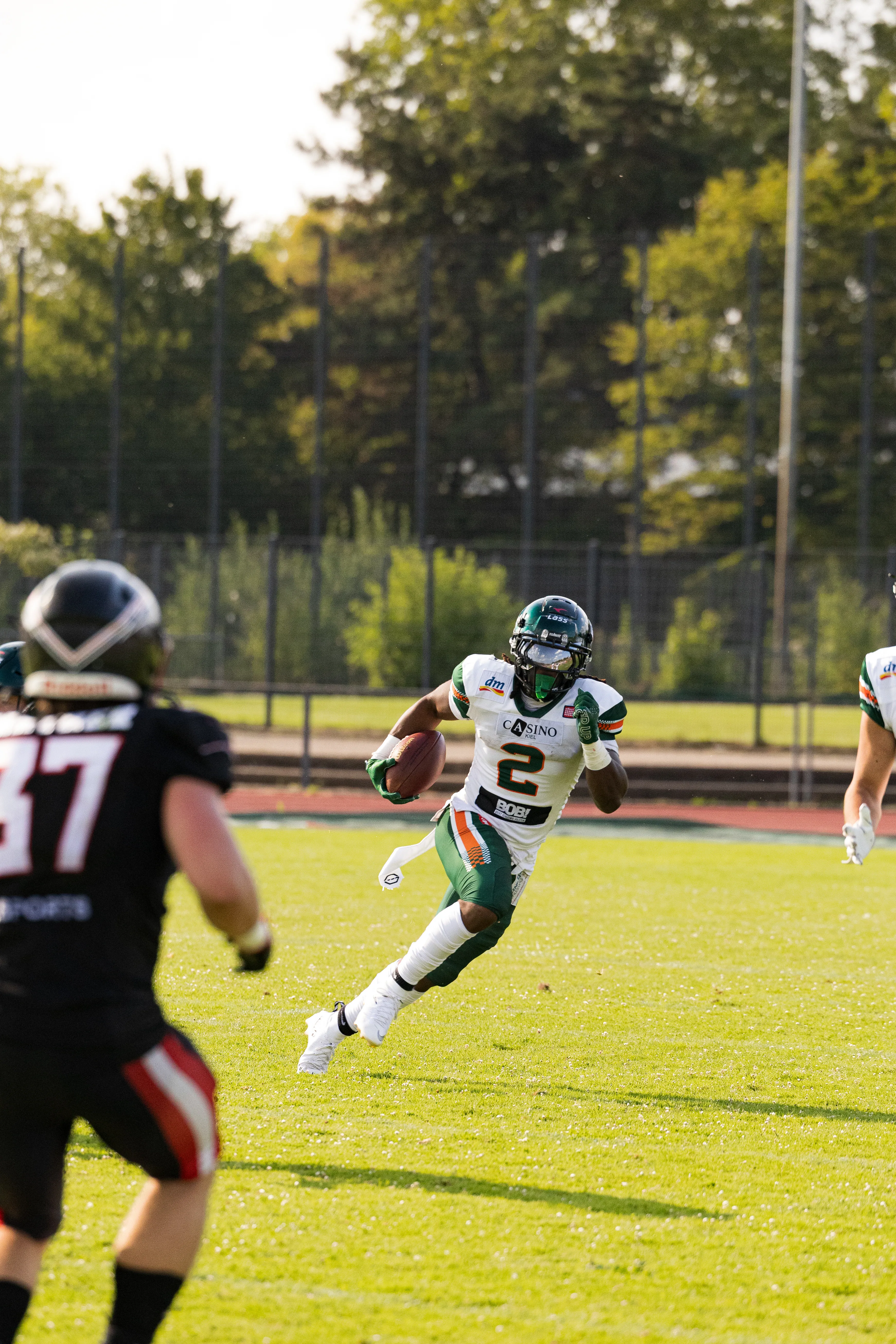 Ein Football-Spieler in einem weißen Trikot mit der Nummer 2 rennt dynamisch mit dem Ball über den Rasen, während sich von links ein gegnerischer Spieler im schwarzen Trikot nähert. Im Hintergrund sind grüne Bäume zu sehen.