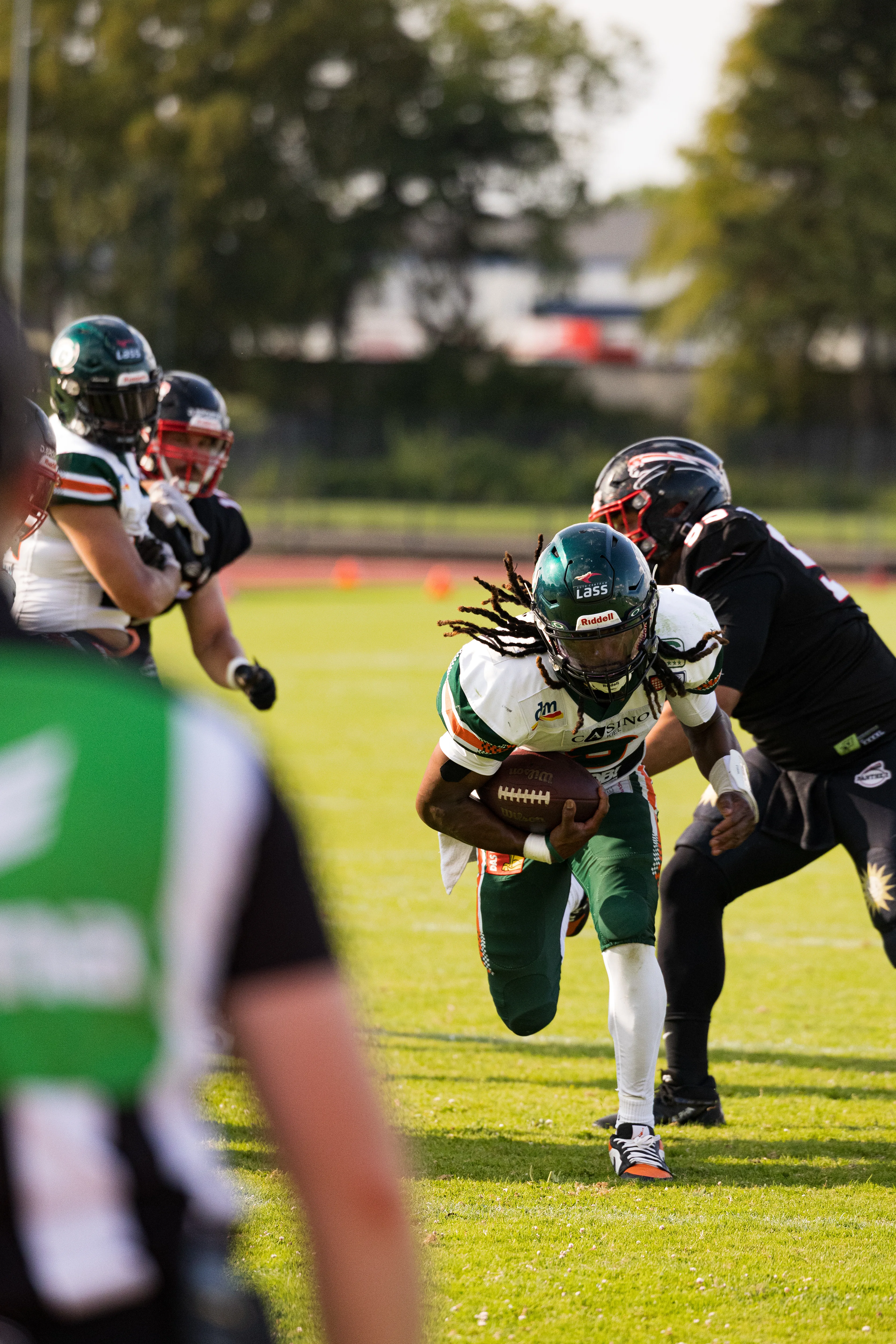 Eine actionreiche Nahaufnahme eines rennenden Football-Spielers mit Dreadlocks im weißen Trikot, der den Ball fest unter dem Arm hält, während ihn ein kräftiger gegnerischer Spieler im schwarzen Trikot verfolgt.